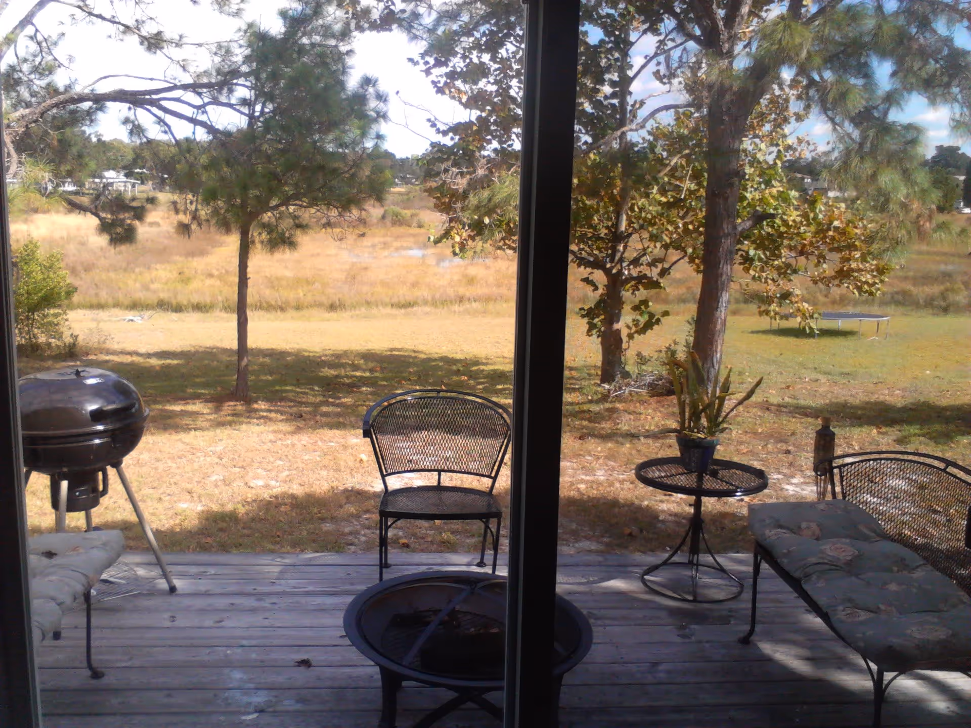 View through a glass door of a wooden deck with outdoor furniture including a cushioned bench, a metal chair, a small round table with a potted plant, a charcoal grill, and a fire pit. Beyond the deck is a grassy yard with trees and a trampoline in the distance under a partly cloudy sky.