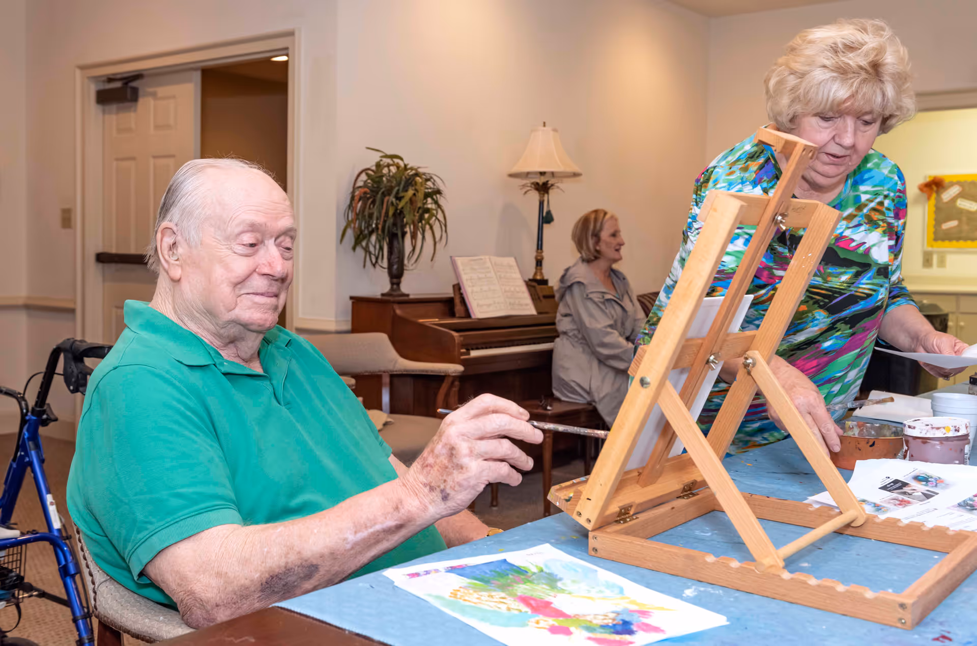 An elderly man in a green shirt is painting at a table with a wooden easel in front of him. A woman in a colorful blouse is standing nearby, assisting or observing. In the background, a woman is sitting at a piano with sheet music, and there is a lamp and a plant on a wooden cabinet.