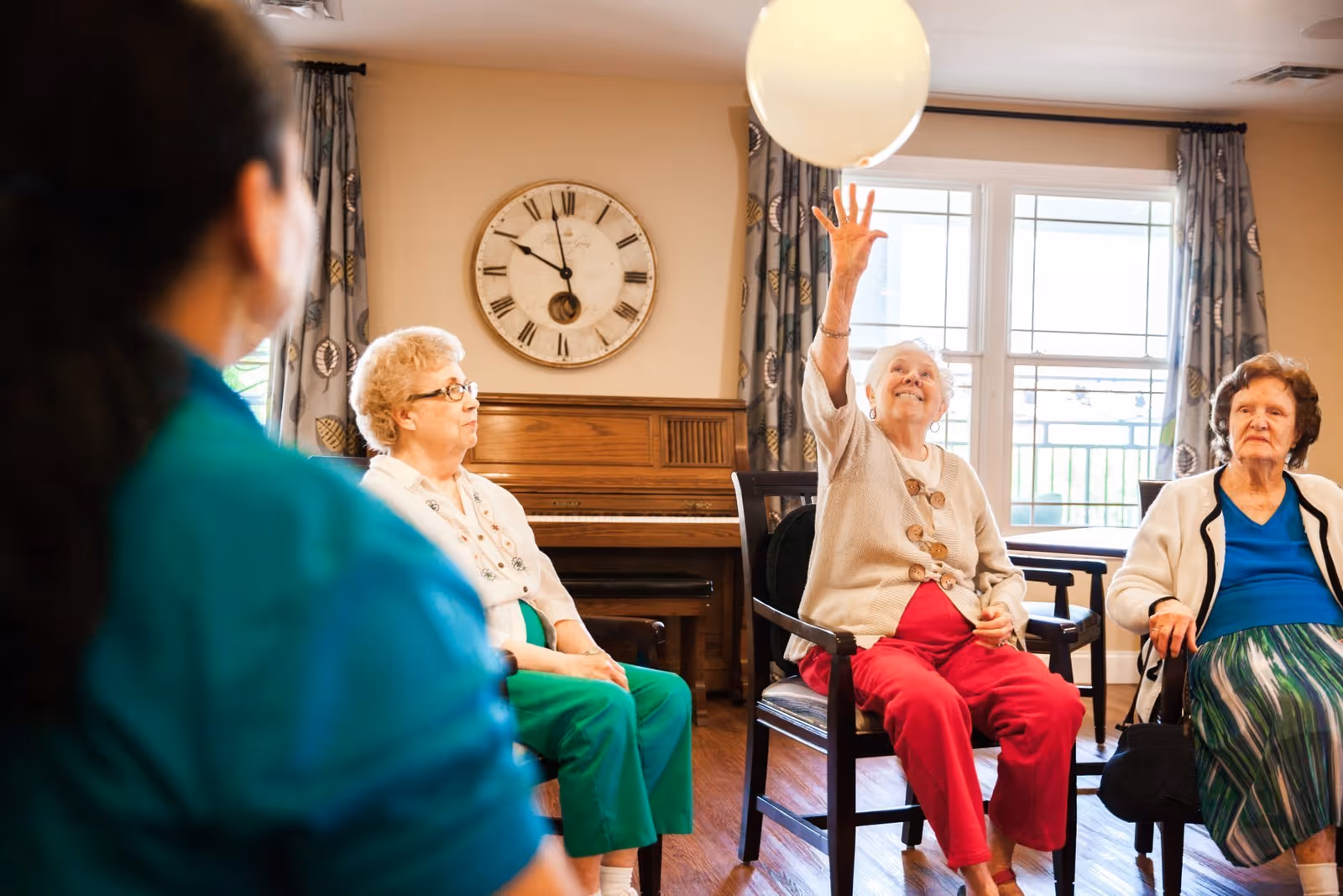 A group of elderly women sitting in chairs in a well-lit room with wooden floors and a piano in the background. One woman is reaching up to catch or hit a white balloon, while the others watch. A large clock is mounted on the wall behind them.