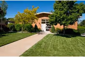 Exterior view of a single-story brick building with large windows and a glass door entrance, surrounded by green grass, trees, and shrubs under a clear blue sky.