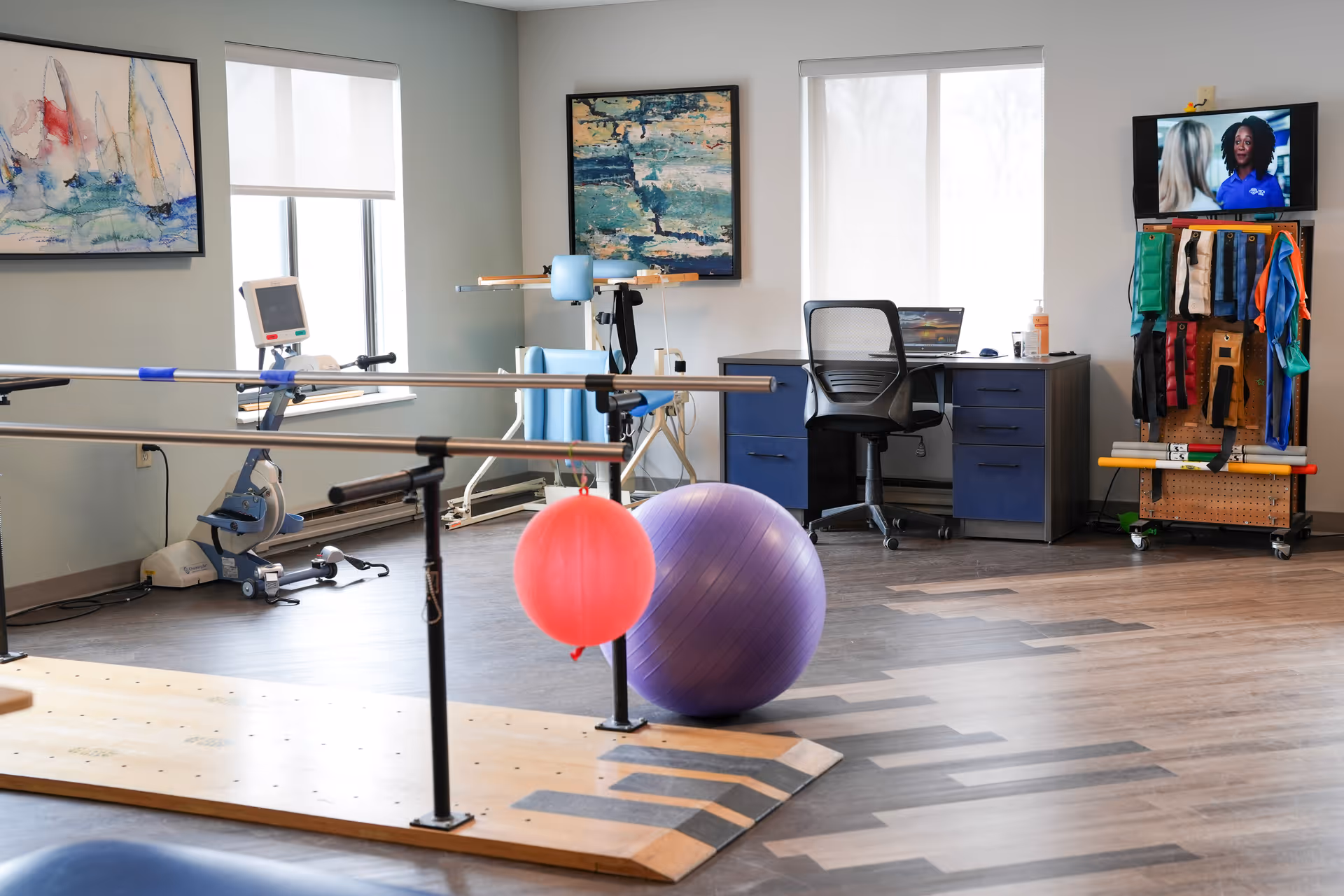 A physical therapy or rehabilitation room with parallel bars for walking exercises, a red exercise ball hanging from the bars, a large purple exercise ball on the floor, a stationary exercise bike, a desk with a chair and laptop, and a TV mounted on the wall showing two people talking. The room has two windows with white blinds and abstract paintings on the walls.