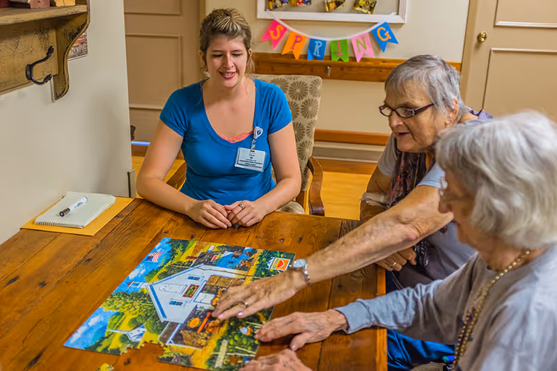 A young female caregiver and two elderly women sitting around a wooden table working together on assembling a colorful jigsaw puzzle in a cozy room with a 'SPRING' banner on the wall.