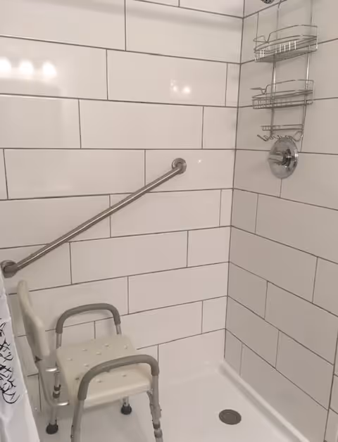 Shower area with white rectangular tiles, a metal grab bar on the wall, a white shower chair with armrests, and a metal shower caddy hanging from the showerhead.