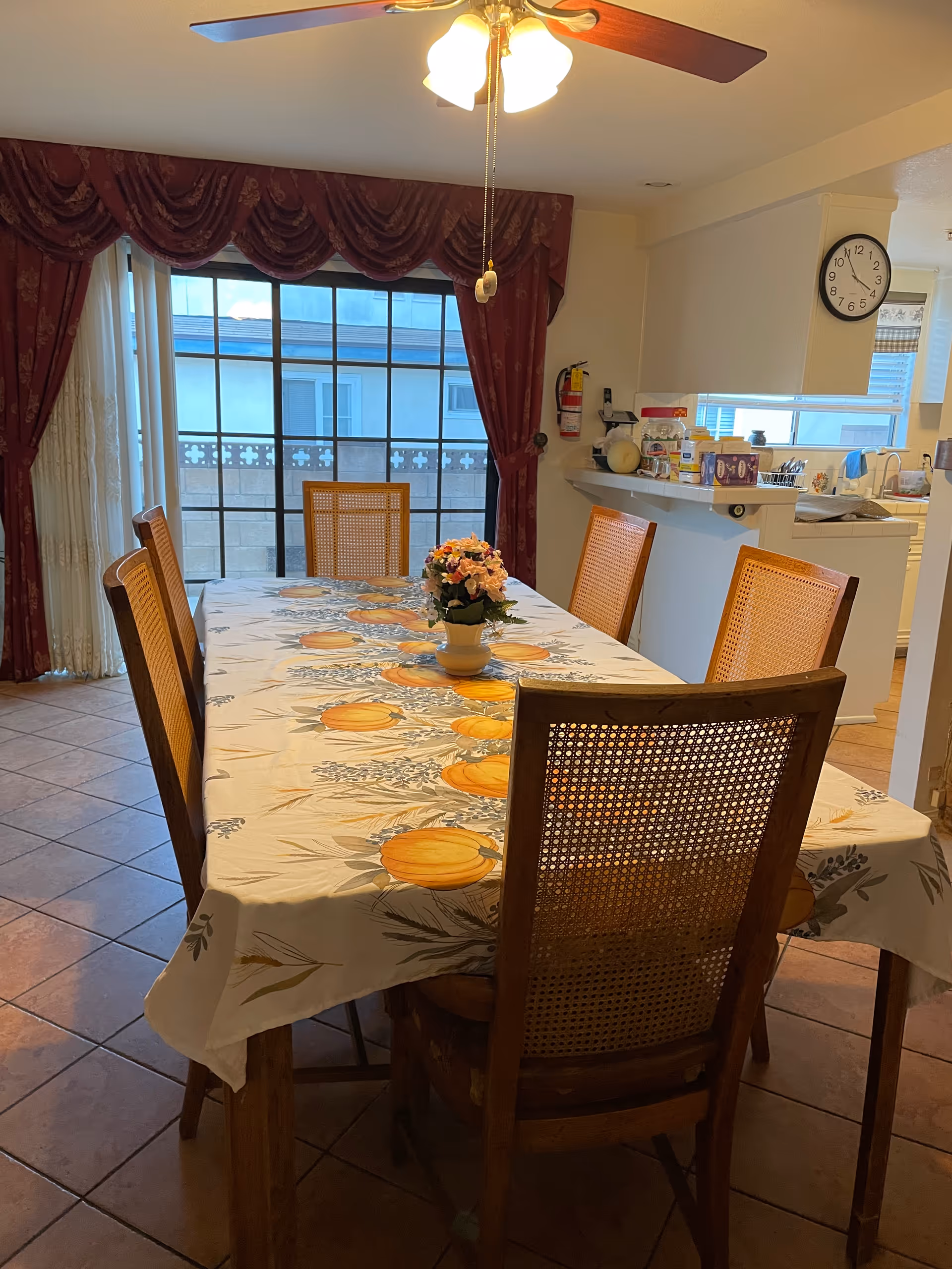 Dining room with a rectangular wooden table covered with a tablecloth featuring pumpkin designs. Six wooden chairs with woven backs surround the table. A small flower arrangement is placed in the center of the table. Behind the table, there is a large window with red curtains and sheer white drapes. To the right, a kitchen counter with various items and a wall clock is visible. The floor is tiled, and a ceiling fan with lights hangs above the table.