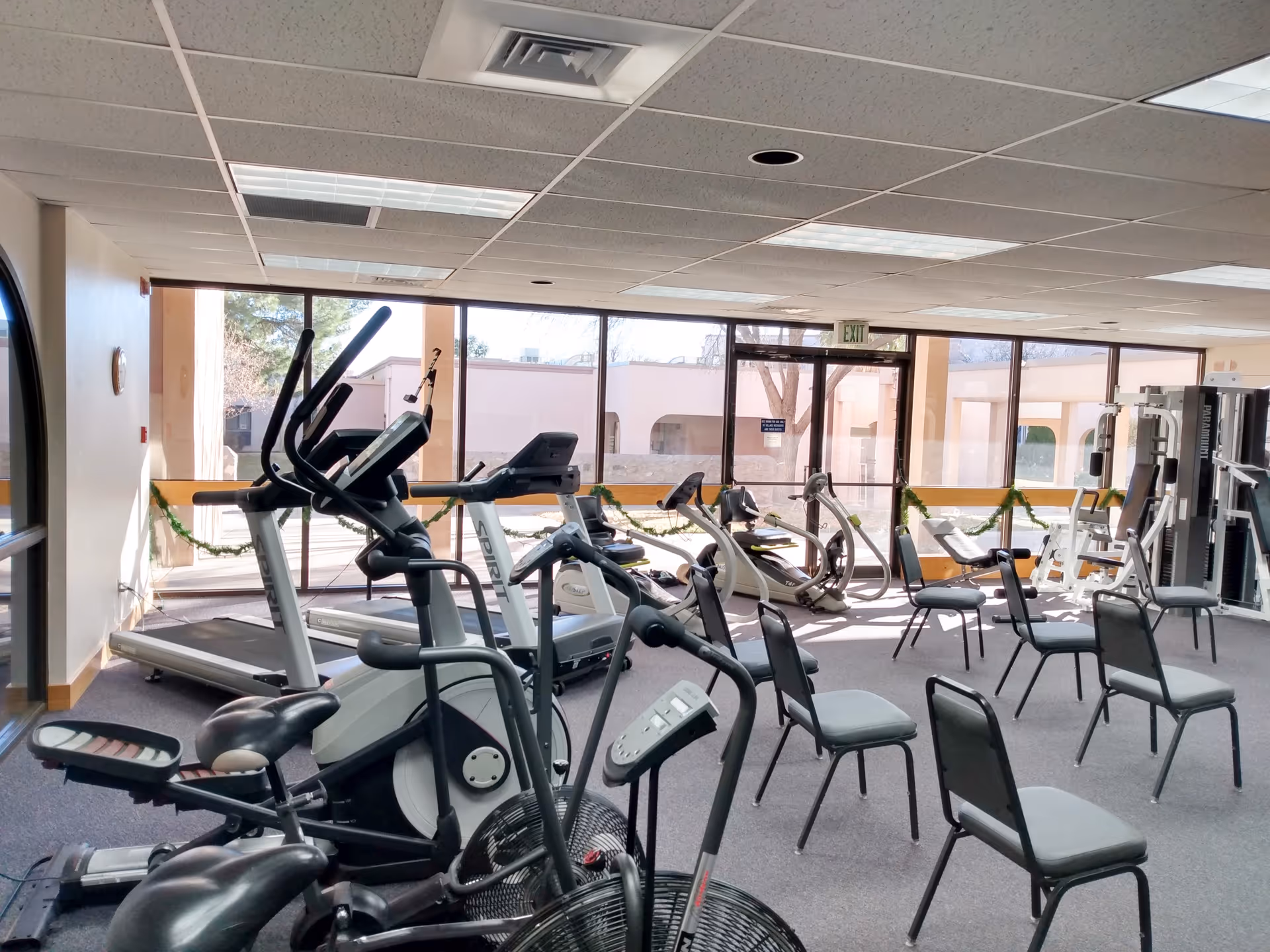 Exercise room with treadmills, ellipticals, stationary bikes, weight machines and chairs by large windows.