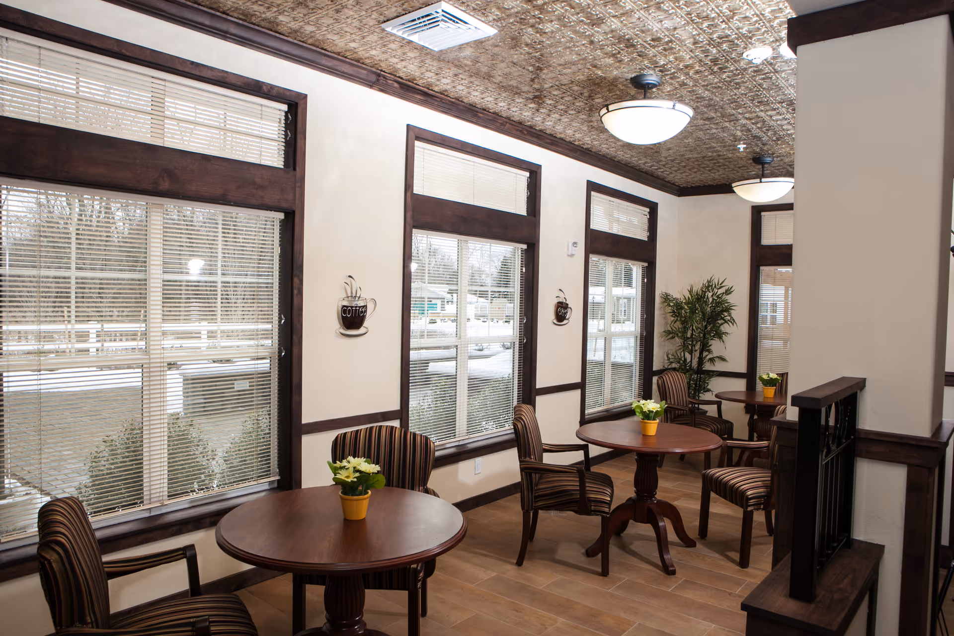 A cozy interior seating area with round wooden tables and striped cushioned chairs. The room has large windows with blinds, decorative wall art shaped like coffee cups, potted plants on the tables and in the corner, and a patterned ceiling with round light fixtures.