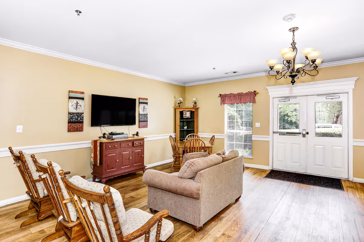 A cozy living room area with beige walls and wooden flooring. The room features a beige sofa, three wooden rocking chairs with cushions, a wooden cabinet with a flat-screen TV mounted above it, and a small round wooden dining table with chairs in the corner. There is a window with red checkered valance and double white doors leading outside. A decorative chandelier hangs from the ceiling.