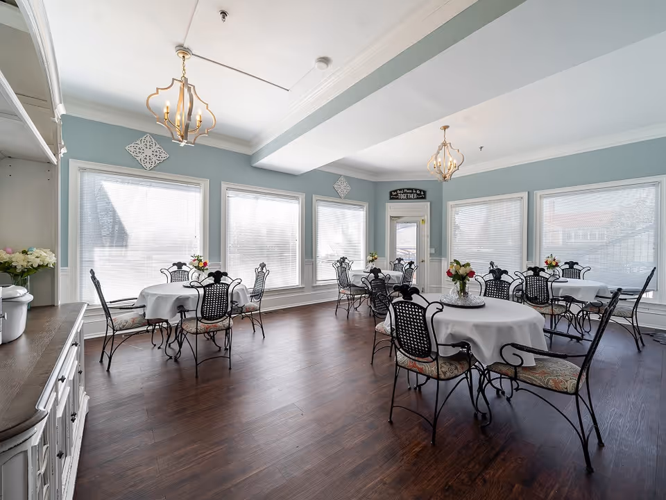 A bright dining room with multiple round tables covered with white tablecloths, each surrounded by black wrought iron chairs with patterned cushions. The room has light blue walls, large windows with blinds, two decorative chandeliers, and floral centerpieces on each table. There is a white sideboard on the left side and a door at the far end with a sign above it that reads 'Our Place Is Better TOGETHER'.