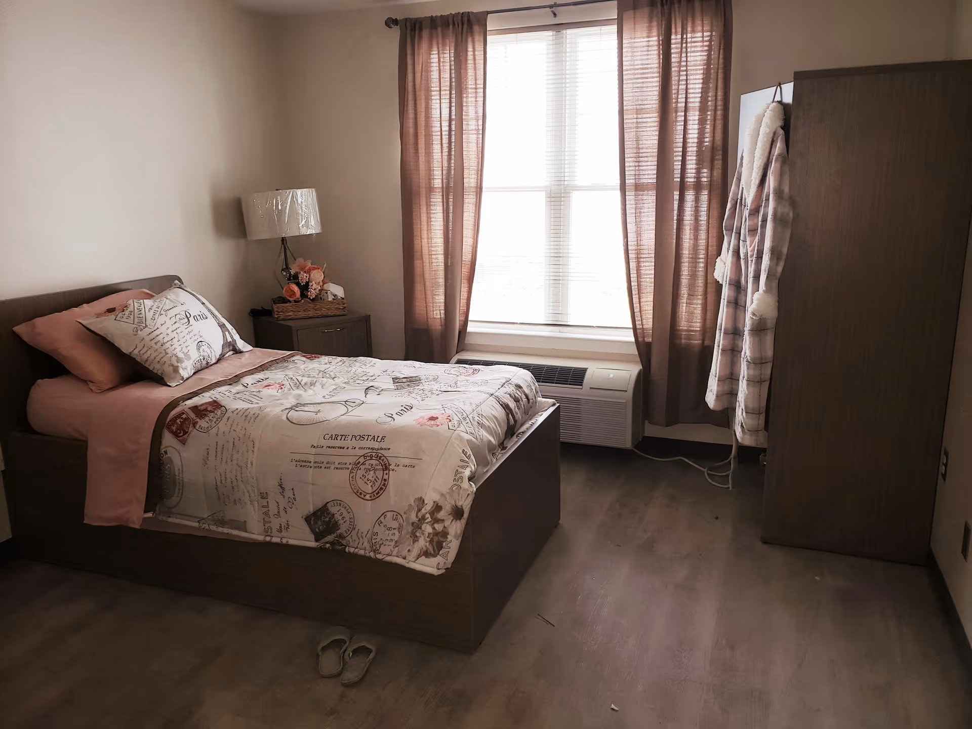 A cozy bedroom with a single bed covered in a Paris-themed comforter and pink sheets. Next to the bed is a small nightstand with a lamp and a basket of flowers. A window with brown curtains lets in natural light. A wardrobe stands in the corner with a plaid robe hanging on its side. A pair of slippers is placed on the wooden floor near the bed.