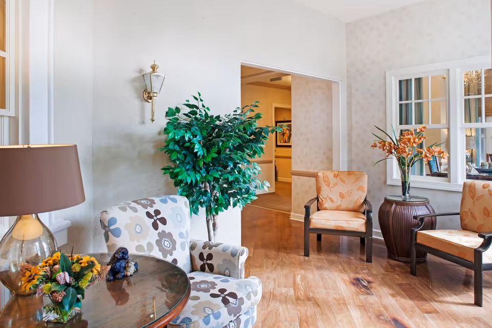 A cozy sitting area in a senior living facility with floral-patterned armchairs, a round wooden table with a lamp and flower arrangement, a large green potted plant, and a side table with a vase of orange flowers. The room has wooden flooring and light-colored walls with a window looking into another room.