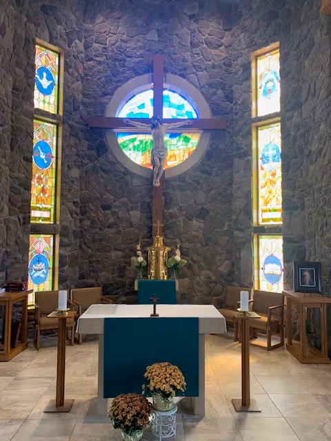 Interior view of a chapel with stone walls, stained glass windows, a large wooden crucifix with a figure of Jesus mounted on the wall, an altar covered with a white cloth and green front panel, two candles on stands, flower arrangements, and chairs arranged around the altar.