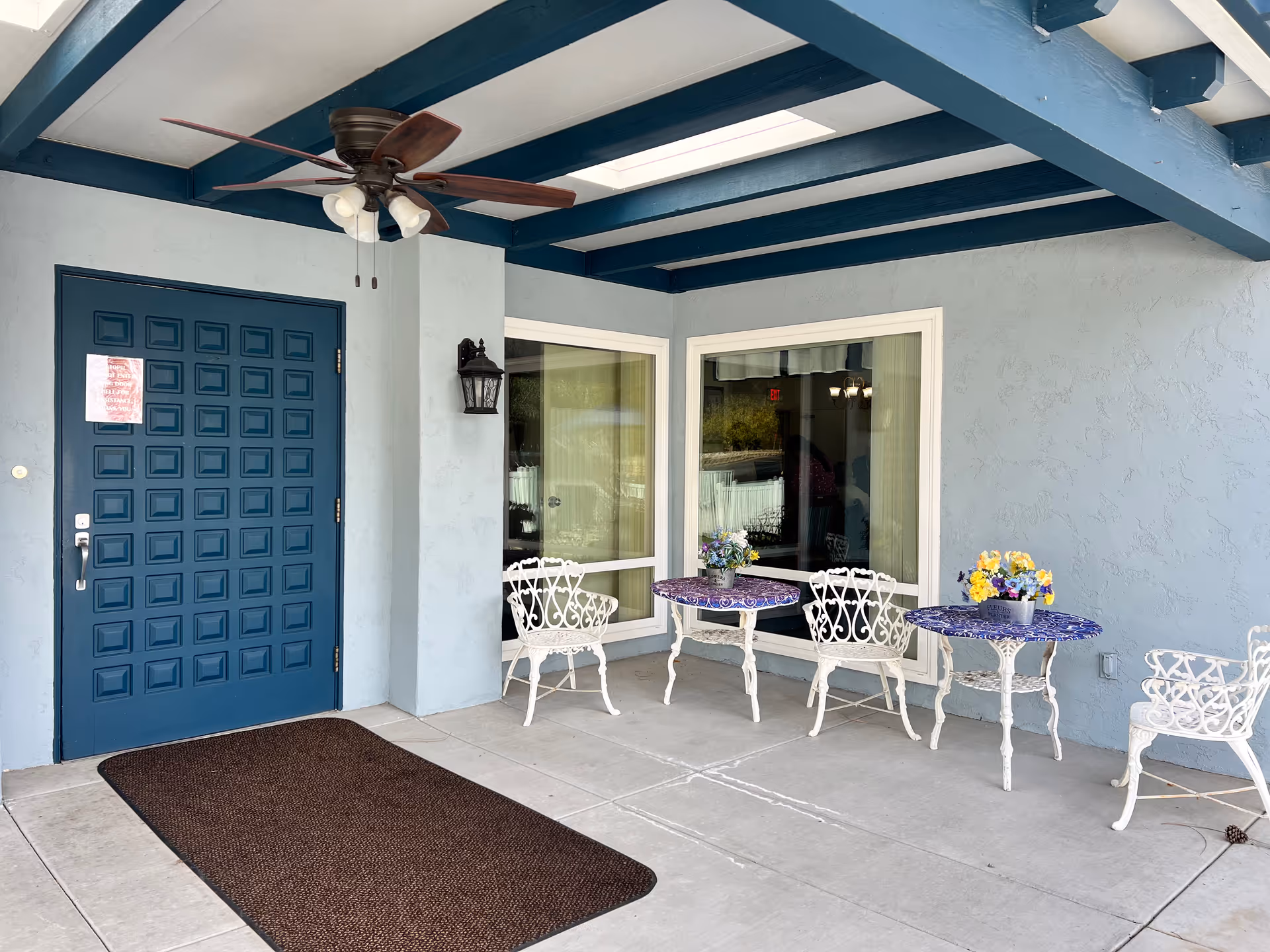 Outdoor covered patio area with a blue door, two white metal tables with purple patterned tops, white metal chairs, flower pots on the tables, a ceiling fan with lights, and large windows on a light blue wall.