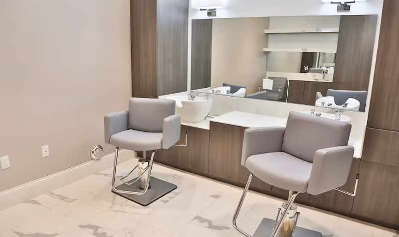 Two gray salon chairs in front of a mirrored grooming station with sinks in a clean modern room.