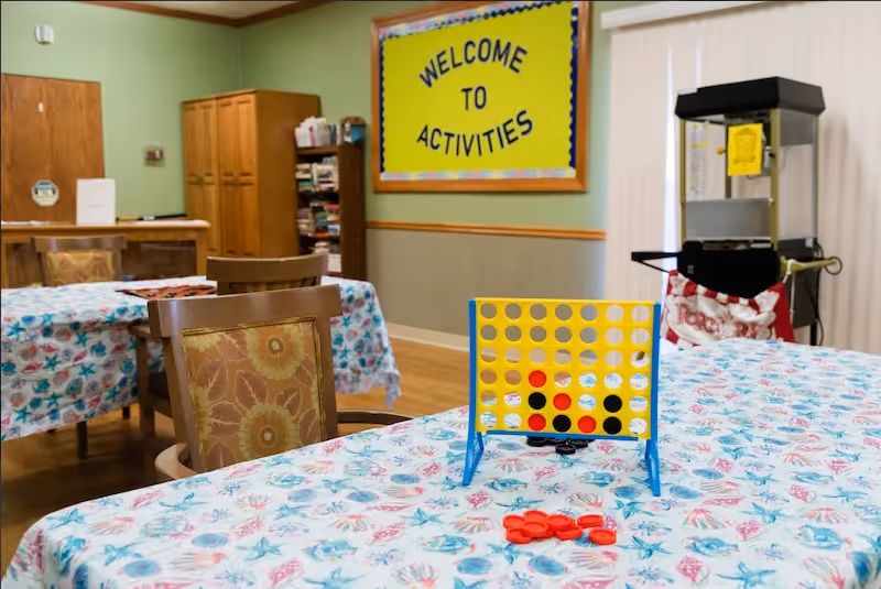 A room with tables covered in floral tablecloths, chairs with patterned cushions, a Connect Four game set up on one table, a popcorn machine in the corner, and a yellow bulletin board on the wall that says 'Welcome to Activities'.