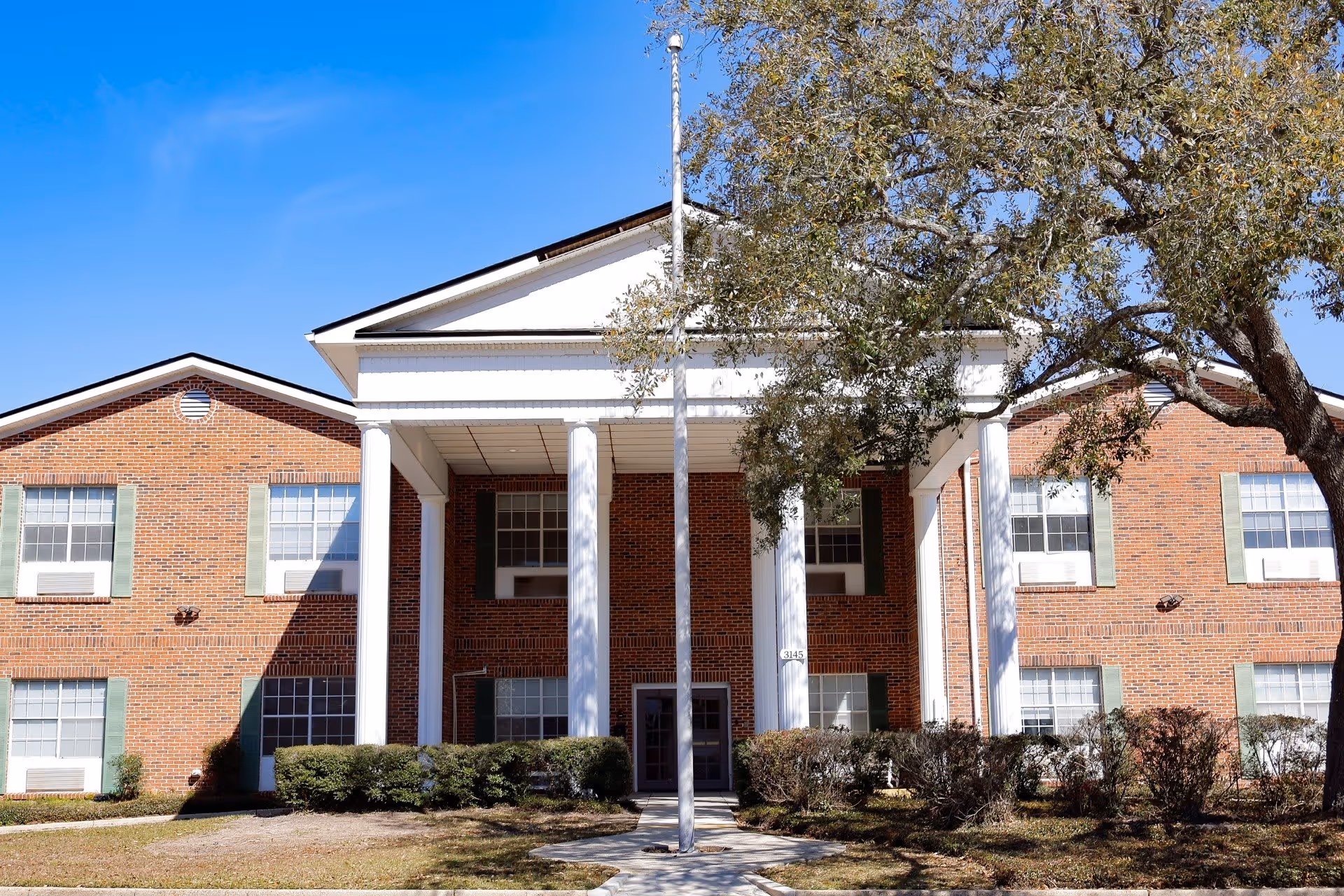Front exterior view of a two-story brick building with white columns and green shutters, surrounded by bushes and a large tree on the right side under a clear blue sky.