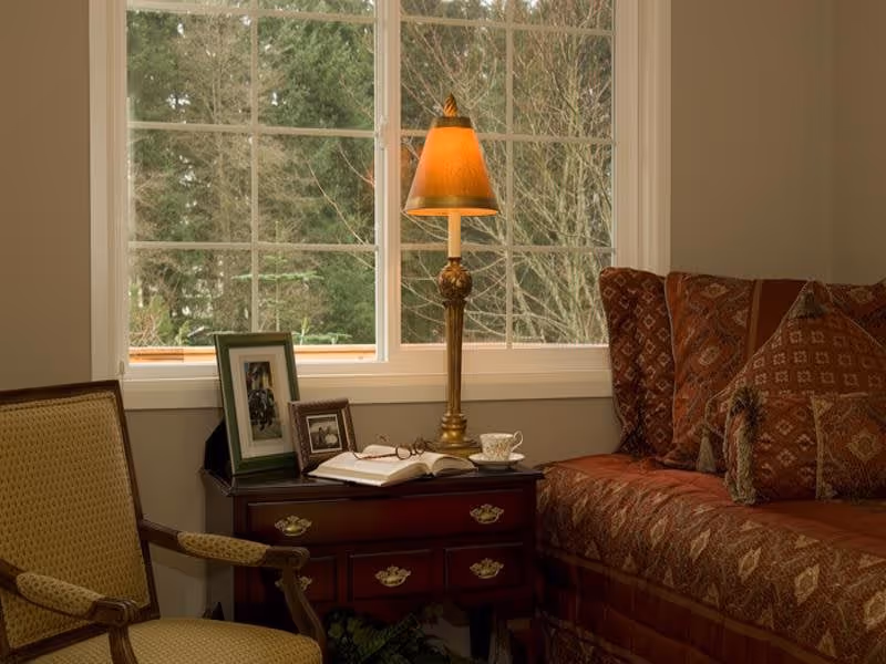 Cozy living room corner with a patterned sofa, armchair, and a wooden side table holding a lamp, open book, teacup, and framed photos in front of a window.