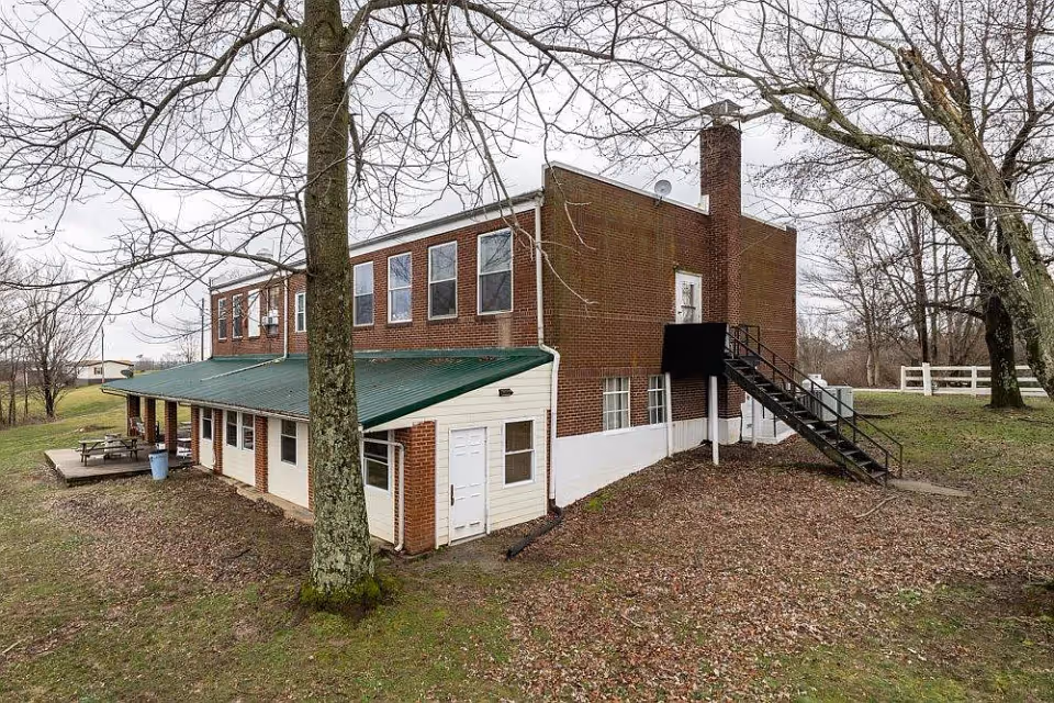 Exterior view of a two-story brick building with a green metal roof over a lower level extension. There are several windows on both floors, a black metal staircase leading to a door on the upper floor, and leafless trees surrounding the building. The ground is covered with fallen leaves and grass.