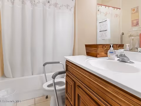 Bathroom with a white shower curtain, a toilet with a gray safety frame around it, a wooden vanity with a white countertop, a sink, a soap dispenser, and a mirror above the sink.