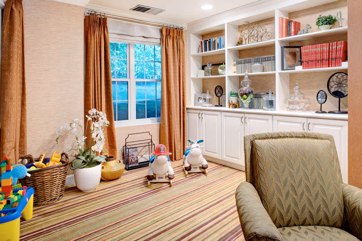 A cozy room with a striped carpet, a large window with brown curtains, and built-in white shelves filled with books and decorative items. There are two rocking horse toys on the floor, a basket with toys, and a green patterned armchair in the foreground.