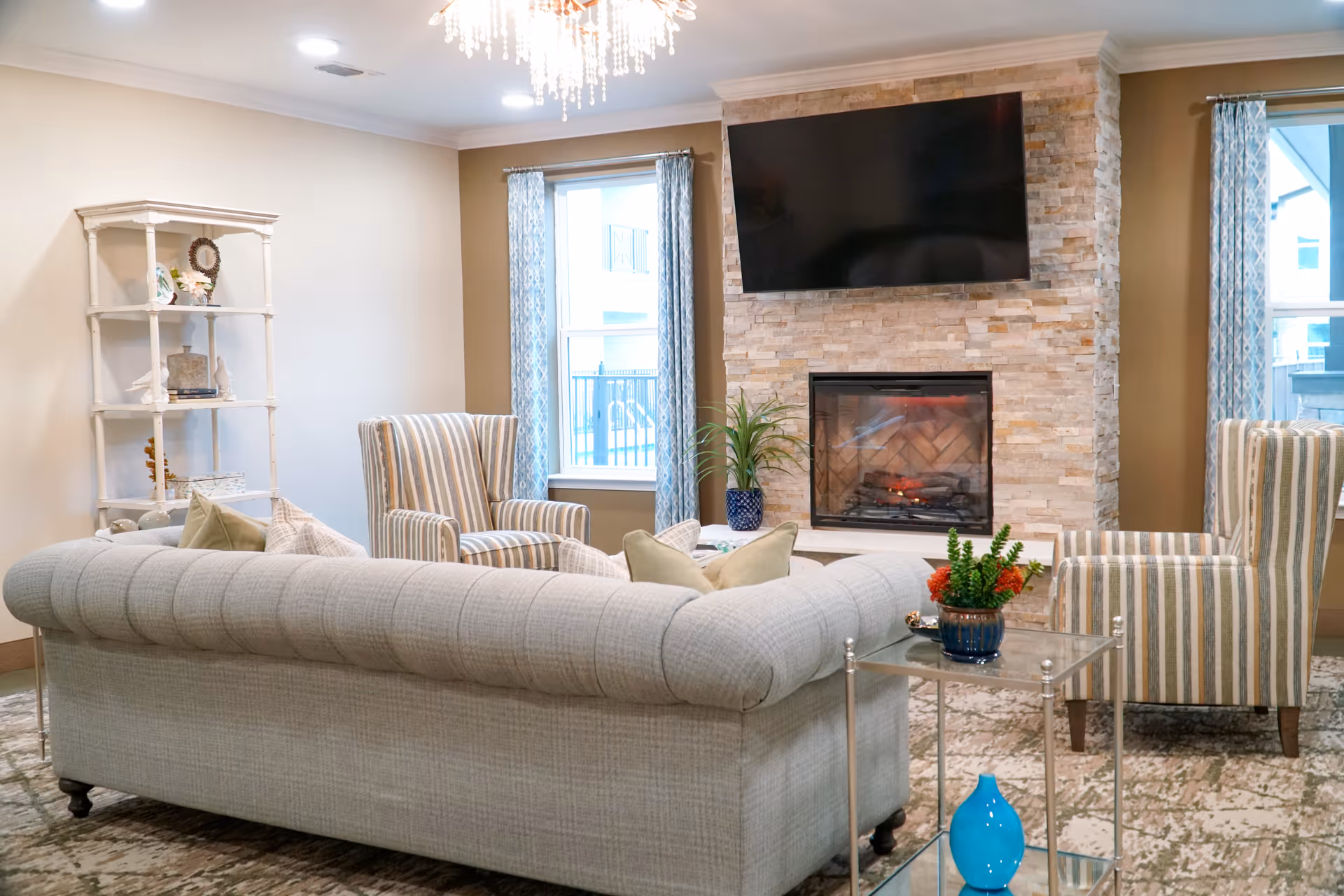 A cozy living room with a tufted light gray sofa, two striped armchairs, a stone fireplace with a mounted flat-screen TV above it, two windows with blue patterned curtains, a white shelving unit with decorative items, and a glass side table with a blue vase and a small potted plant.