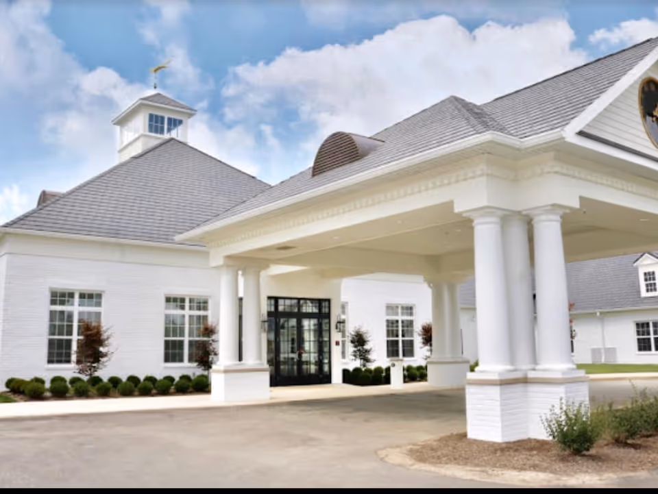 Front exterior view of a white senior living facility building with a covered entrance supported by large white columns, neatly trimmed bushes, and a clear blue sky with some clouds.