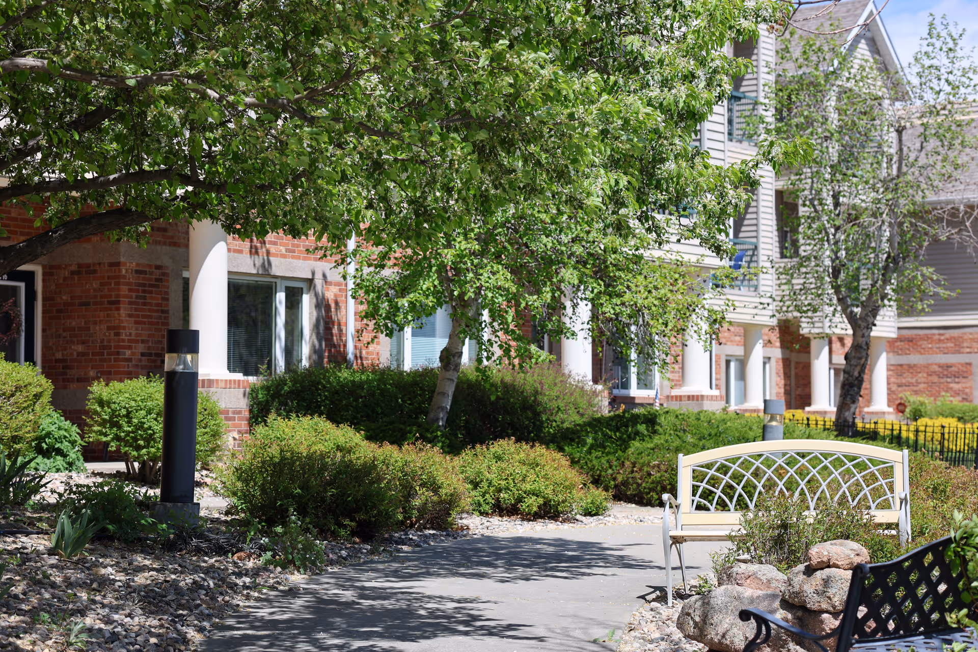 Outdoor garden area at The Wexford facility with a paved walkway, green bushes, trees providing shade, and benches for seating. The building exterior features brick and siding with white columns.