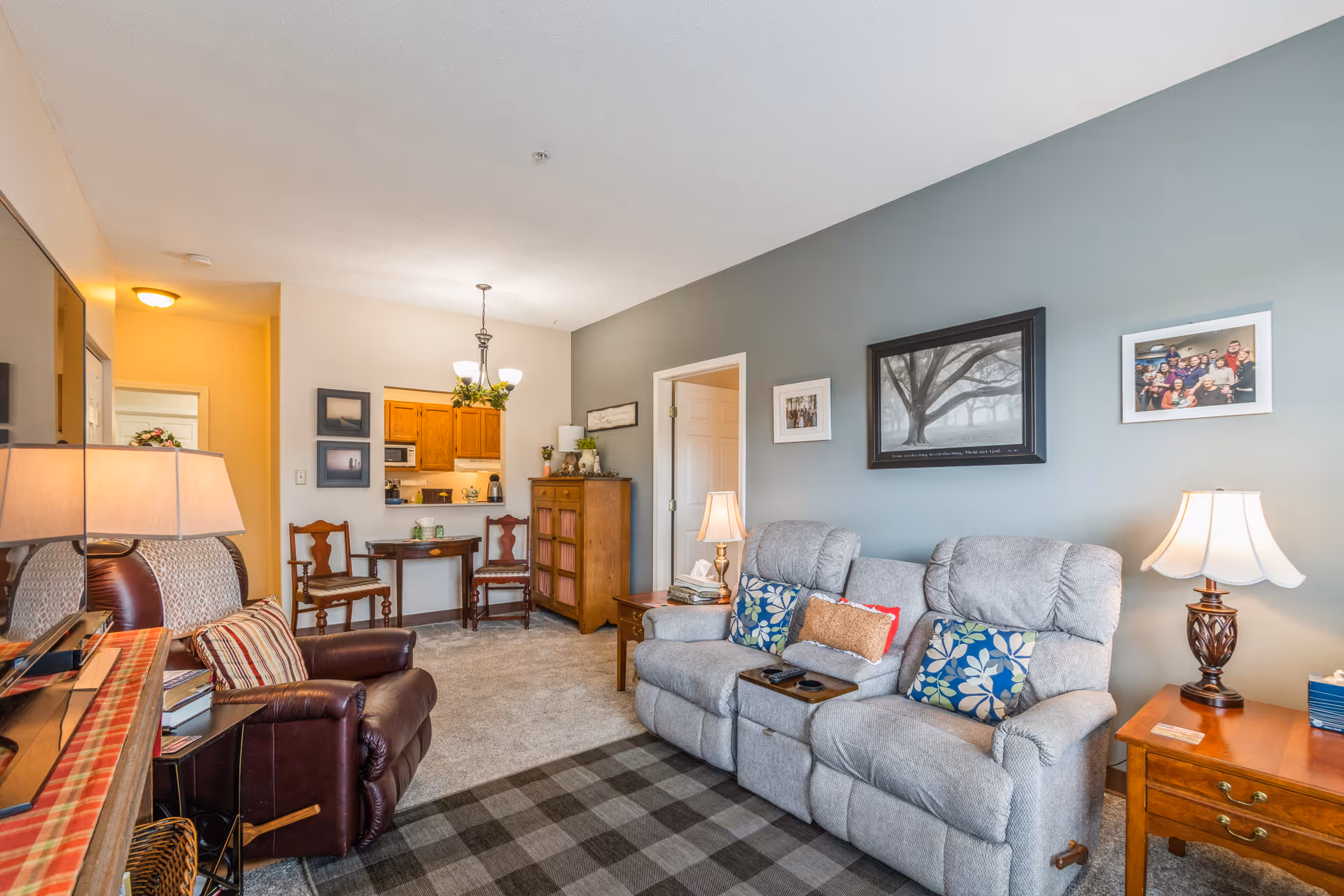 A cozy living room with two gray recliner chairs adorned with floral and solid pillows, a wooden side table with a lamp, and a leather armchair with a striped pillow. The room has a blue-gray accent wall decorated with framed pictures, including a large tree photograph. In the background, there is a small dining area with two wooden chairs and a table, and a kitchen with wooden cabinets and a hanging light fixture.