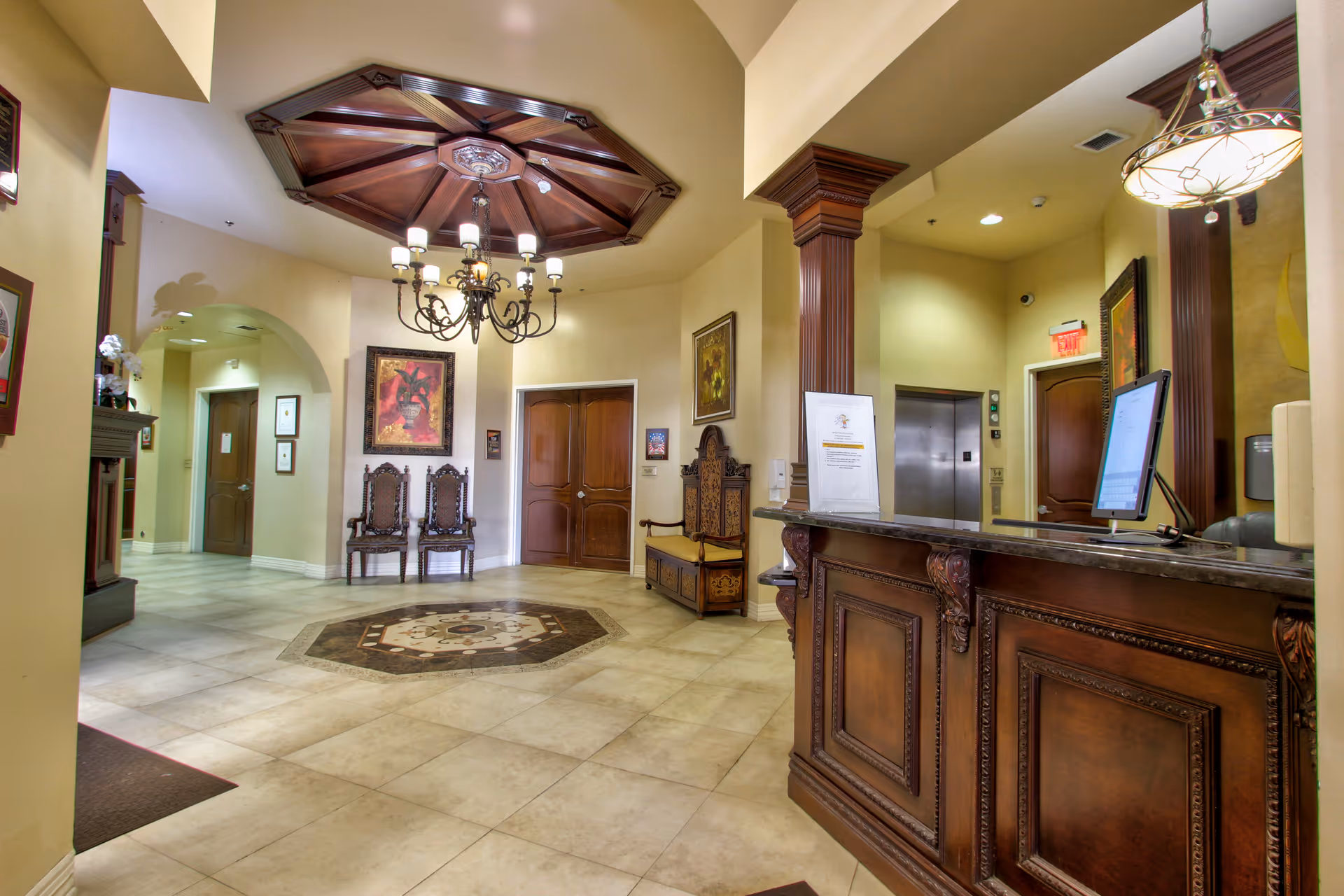 Interior view of a reception area with a wooden front desk, a computer monitor, decorative wooden chairs, framed artwork on the walls, a chandelier hanging from a wooden ceiling feature, and an elevator door in the background.