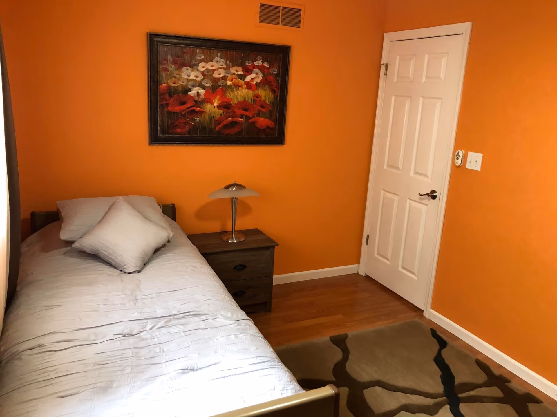 A small bedroom with bright orange walls, a single bed with white bedding and two pillows, a wooden nightstand with a modern lamp, a framed floral painting on the wall, a white door, and a patterned area rug on a wooden floor.