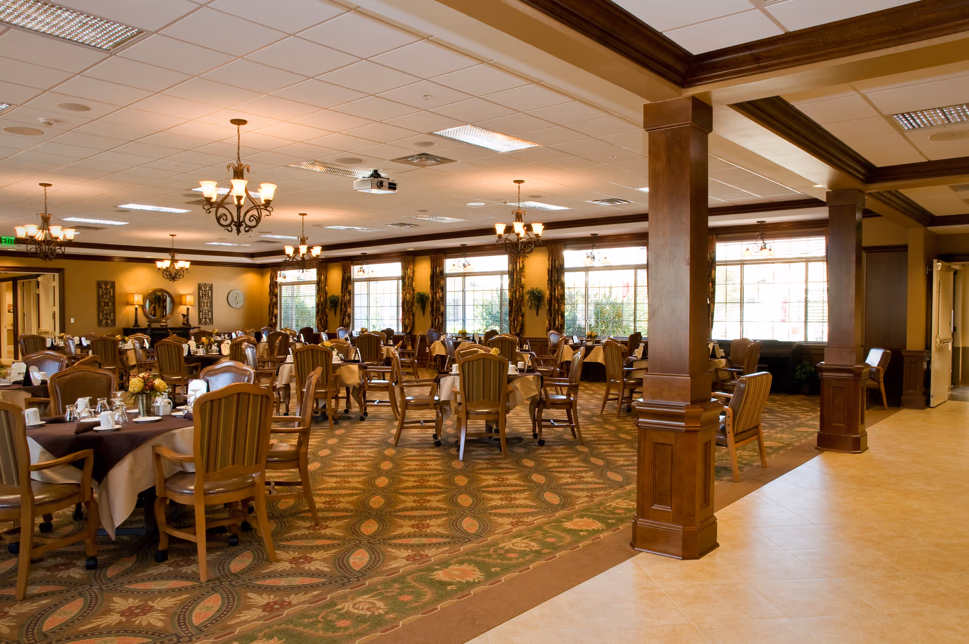 Spacious dining room with round tables set for meals, wooden chairs, chandeliers, and large windows.