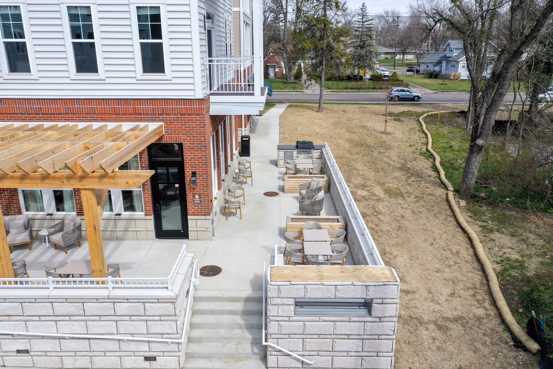 Outdoor patio area at Brookwood Point featuring seating arrangements with chairs and tables, a wooden pergola, and a brick and white siding building. The patio overlooks a grassy area with trees and a road in the background.