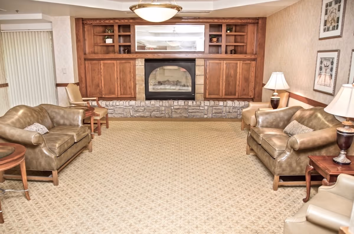 A cozy senior living room with two brown leather sofas facing each other, beige armchairs, wooden side tables with lamps, and a fireplace with a stone base and wooden cabinets above it. The room has beige patterned carpet and light-colored wallpaper with framed artwork on the walls.