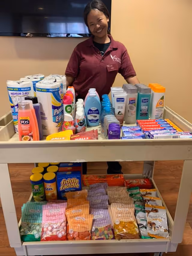 A smiling woman wearing a maroon Victory Centre of River Woods shirt stands behind a cart filled with various personal care products like shampoo, lotion, deodorant, toothpaste, and paper towels on the top shelf, and assorted snacks including candy and chips on the bottom shelf.