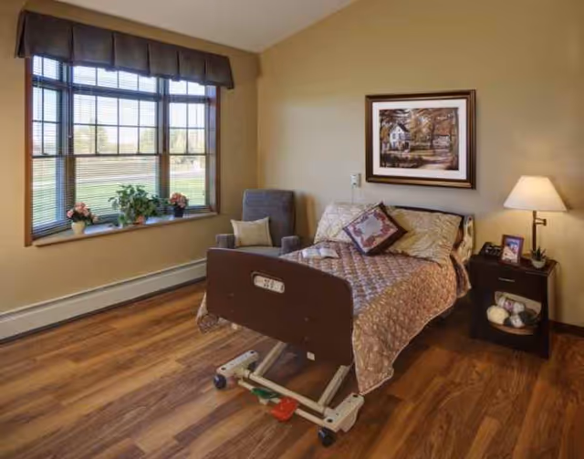 A hospital-style adjustable bed in a cozy resident room with window and nightstand.