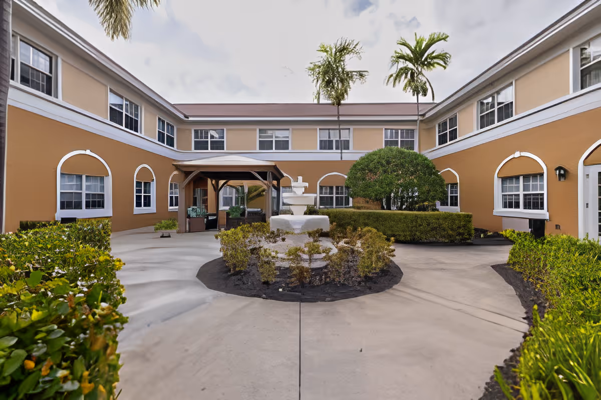 Outdoor courtyard area of a senior living facility with a central white fountain surrounded by small bushes, a covered seating area with chairs, palm trees, and a two-story building with multiple windows and beige walls.