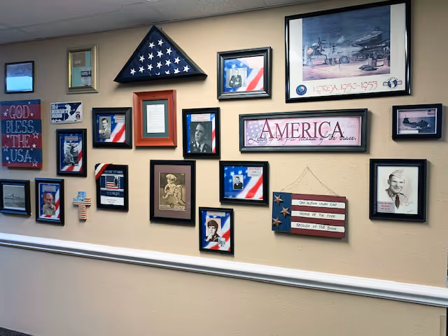 A beige wall decorated with multiple framed patriotic pictures and memorabilia, including American flags, photos of veterans, and signs with patriotic messages such as 'God Bless the USA' and 'America'. There is also a folded American flag in a triangular display case mounted on the wall.