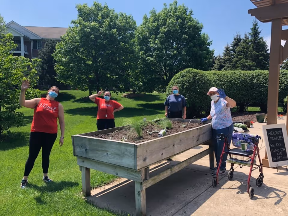 Four people wearing face masks are outdoors around a raised garden bed with some small plants. Three individuals are standing on the grass behind the garden bed, waving, while one person with a walker is standing on the concrete patio next to the garden bed, also waving. There are green trees and bushes in the background, and a sign on the patio reads, 'WELCOME FAMILIES WE ARE GLAD YOU ARE HERE!!!!'.