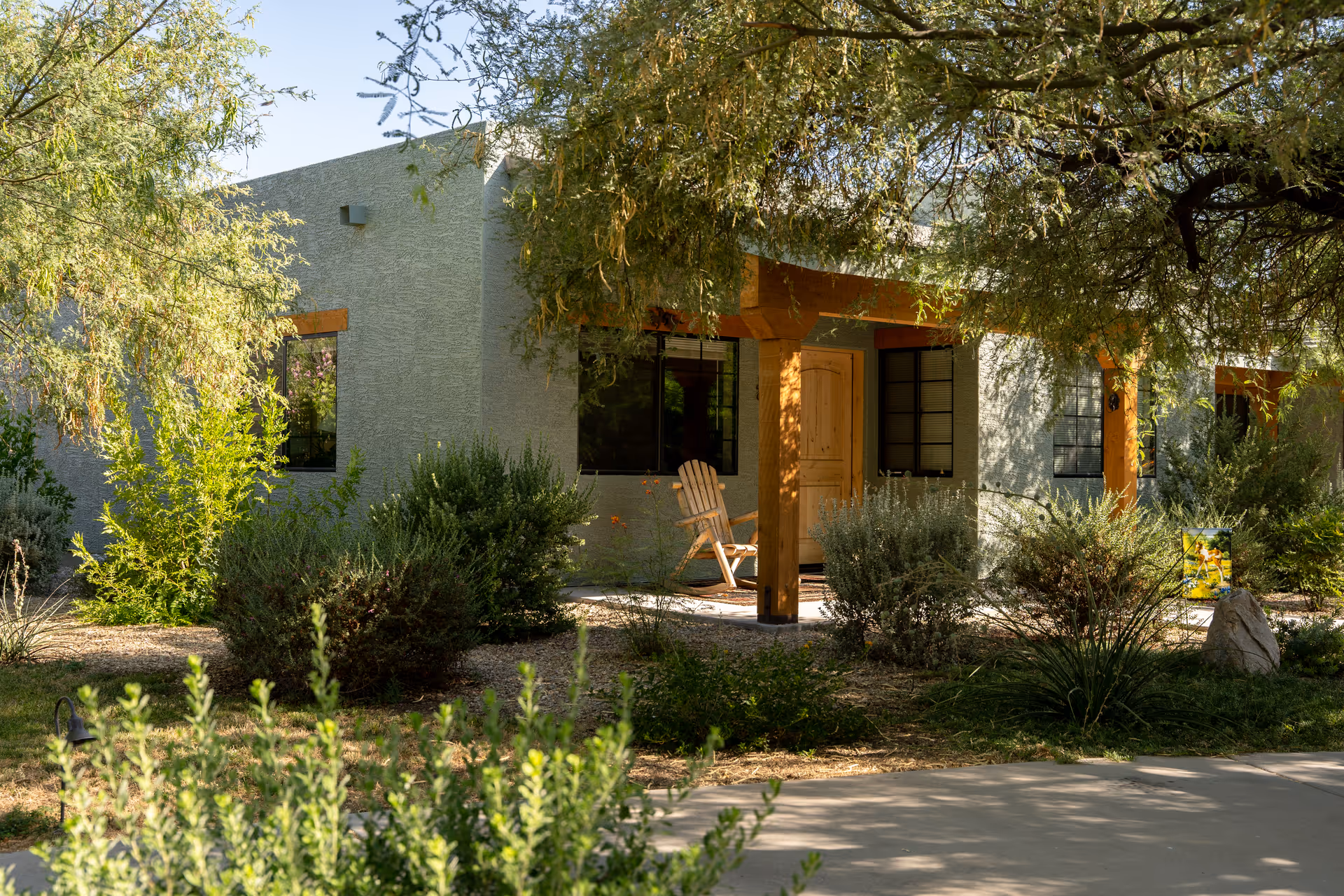 Exterior view of a single-story building with a covered porch supported by wooden beams. There is a wooden chair on the porch, surrounded by various green shrubs and trees in a landscaped garden area under natural sunlight.