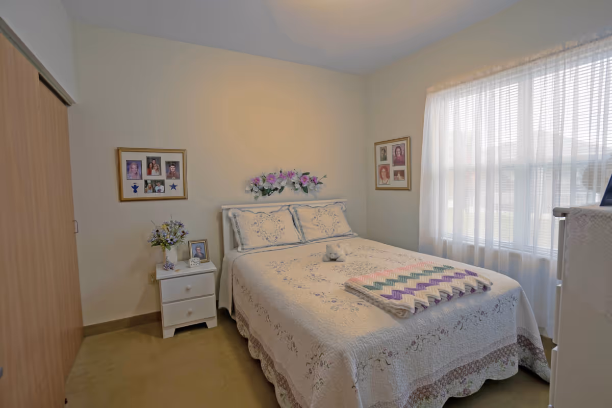 A cozy bedroom with a double bed covered in a white floral quilt and matching pillows. A colorful knitted blanket is folded at the foot of the bed, and a small stuffed animal is placed on the bed. There is a white nightstand with a vase of flowers, a framed photo, and a small clock. Two framed photo collages hang on the walls, and a large window with sheer white curtains allows natural light to fill the room. A wooden closet door is visible on the left side.