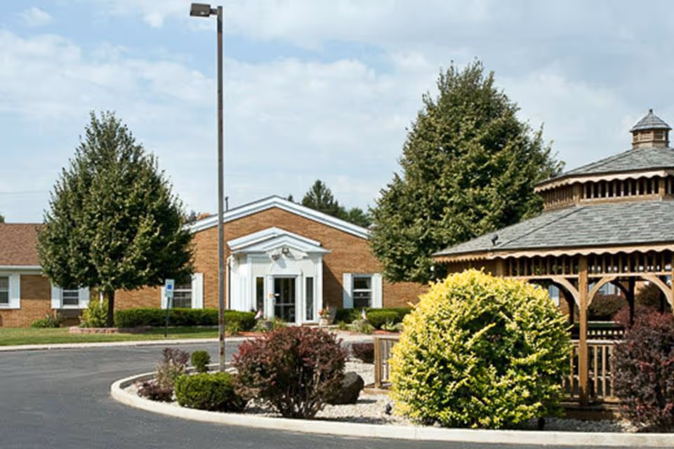 Front exterior of a brick senior living facility with a central entrance, landscaped driveway, and a wooden gazebo.