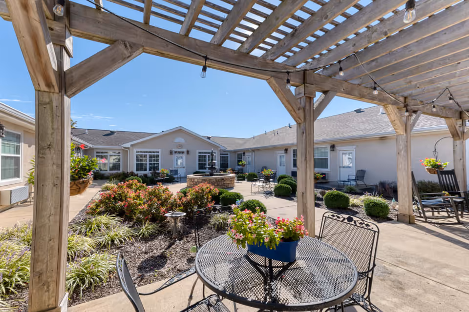 Sunlit courtyard with a wooden pergola, metal patio table and chairs, flower beds and a central fountain surrounded by single-story facility buildings.