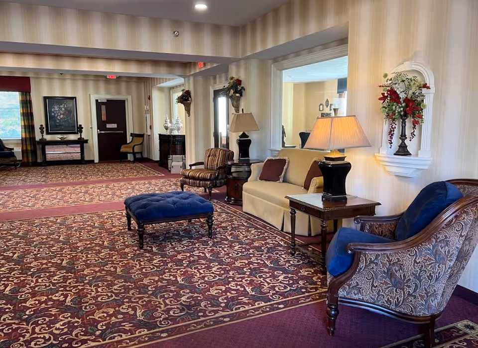 A spacious and elegantly decorated common area with patterned carpet, a beige sofa with two pillows, a blue cushioned armchair, a striped armchair, a blue ottoman, side tables with lamps, floral wall decorations, and a large mirror on the wall. There is a door and window with curtains in the background.