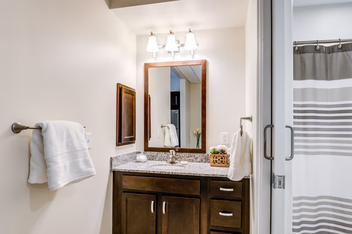 A bathroom vanity with a granite countertop, a rectangular mirror with a wooden frame, and a three-light fixture above. There are white towels hanging on towel racks on either side of the vanity. To the right, a shower curtain with gray and white stripes is partially visible.
