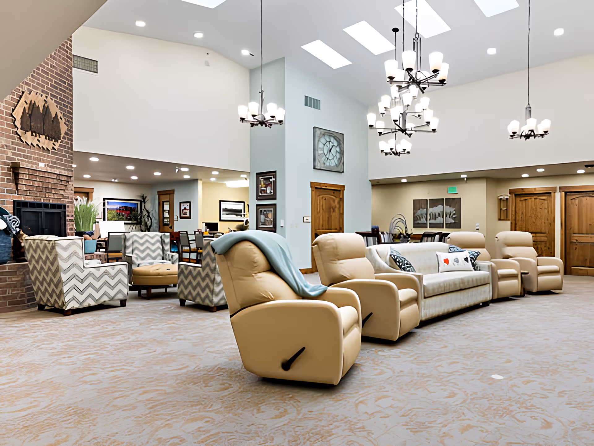 A spacious and well-lit living room area in Mill View Memory Care featuring beige recliner chairs and a matching sofa arranged in a semi-circle. The room has high ceilings with skylights and multiple chandeliers. There is a brick fireplace on the left side with a wooden art piece above it, and additional seating with patterned armchairs and a small ottoman near the fireplace. The walls are decorated with framed pictures and wooden doors are visible in the background.