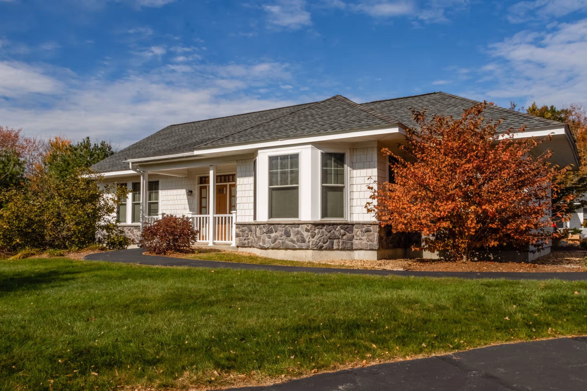 Exterior view of a single-story residential building with white siding, stone accents, and a gray shingled roof. The building is surrounded by green grass, a paved walkway, and trees with autumn foliage under a partly cloudy blue sky.