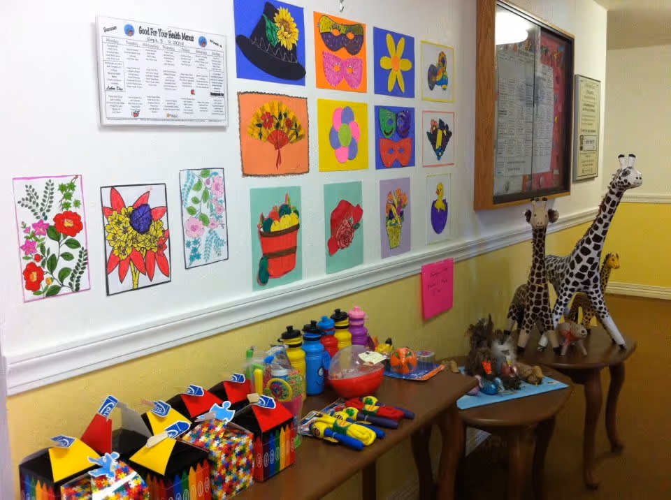 A hallway wall decorated with colorful artwork including flowers, masks, and fruit baskets. Below the artwork, there are tables displaying various items such as colorful water bottles, toys, and stuffed giraffes. The wall is painted yellow on the lower half and white on the upper half, with a bulletin board and a menu posted on the wall.