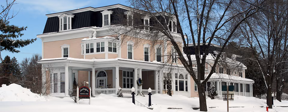 A large, historic-style building with light pink siding and white trim, covered in snow. The building has multiple windows, a black mansard roof, and a wrap-around porch with white columns. Snow-covered trees and a clear blue sky are visible around the building. A sign near the entrance reads 'Redington Memorial Home.'