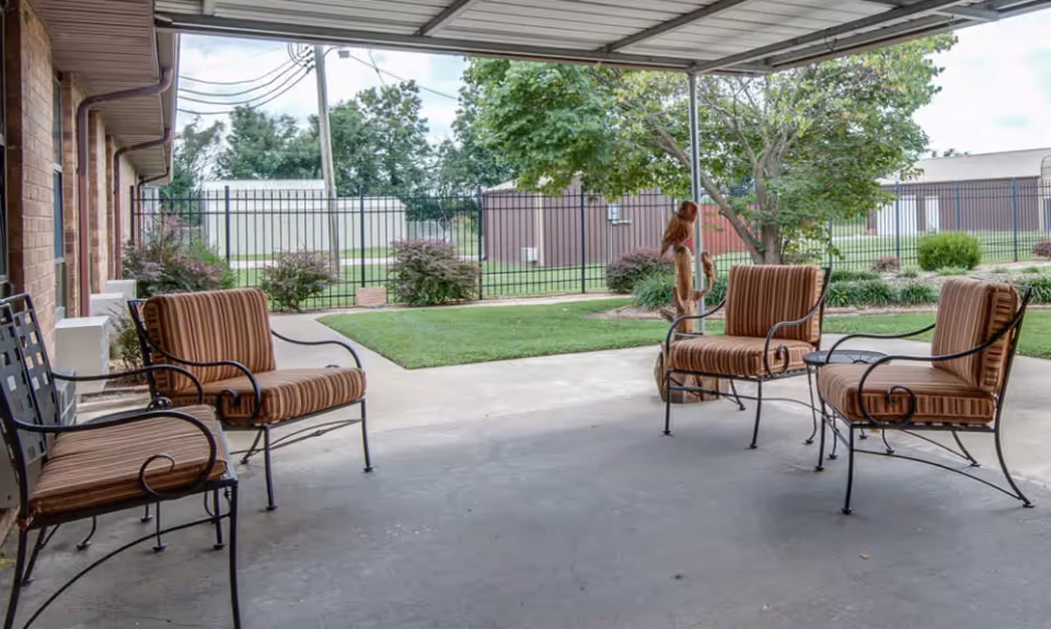 Covered outdoor patio area with four metal chairs featuring brown striped cushions arranged around the space. The patio overlooks a grassy area with bushes, trees, and a black metal fence enclosing the yard. The sky is partly cloudy.