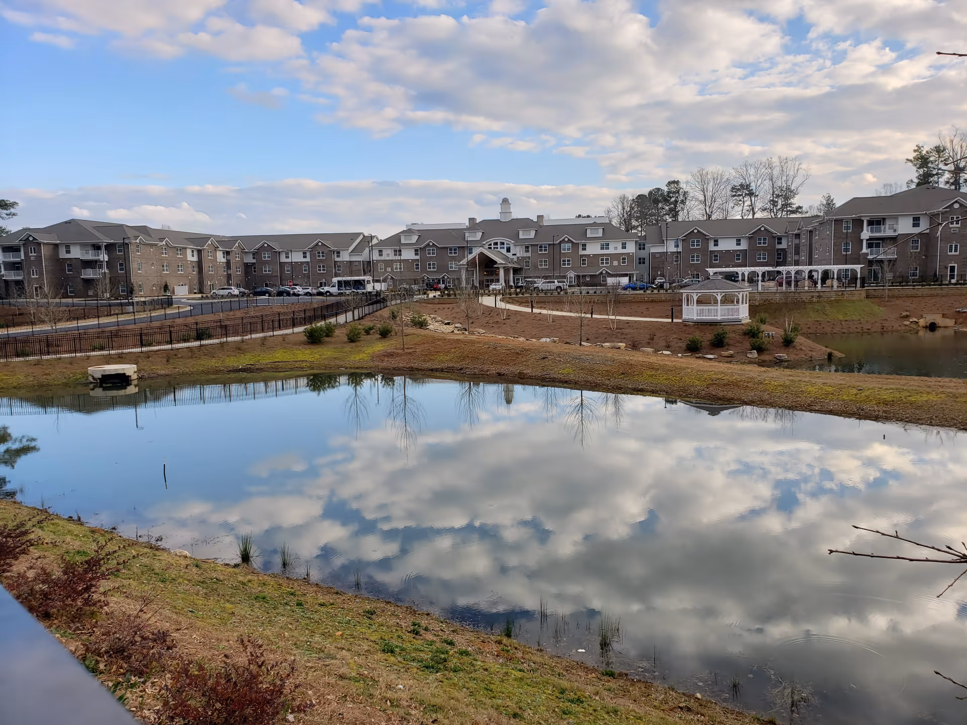 View of Vickery Rose Retirement Resort showing a large multi-story building in the background with a pond reflecting the cloudy sky in the foreground. There is a gazebo near the pond and landscaped grounds with pathways and some small trees and shrubs.