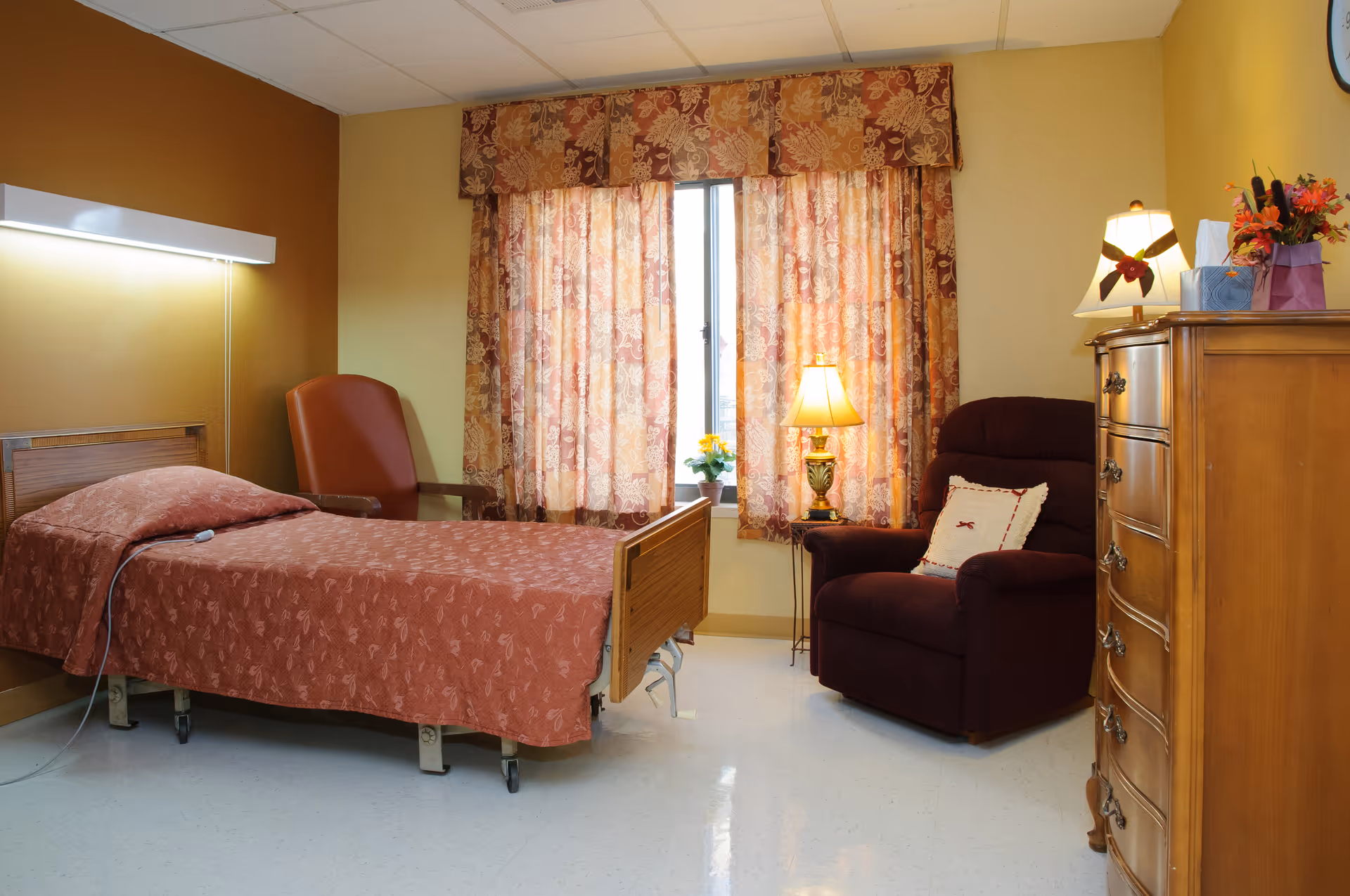 A cozy senior living facility room with a single bed covered in a patterned rust-colored bedspread, a matching armchair, a wooden dresser, and a window with floral curtains. A small table lamp and a flower vase are placed near the window, creating a warm and inviting atmosphere.